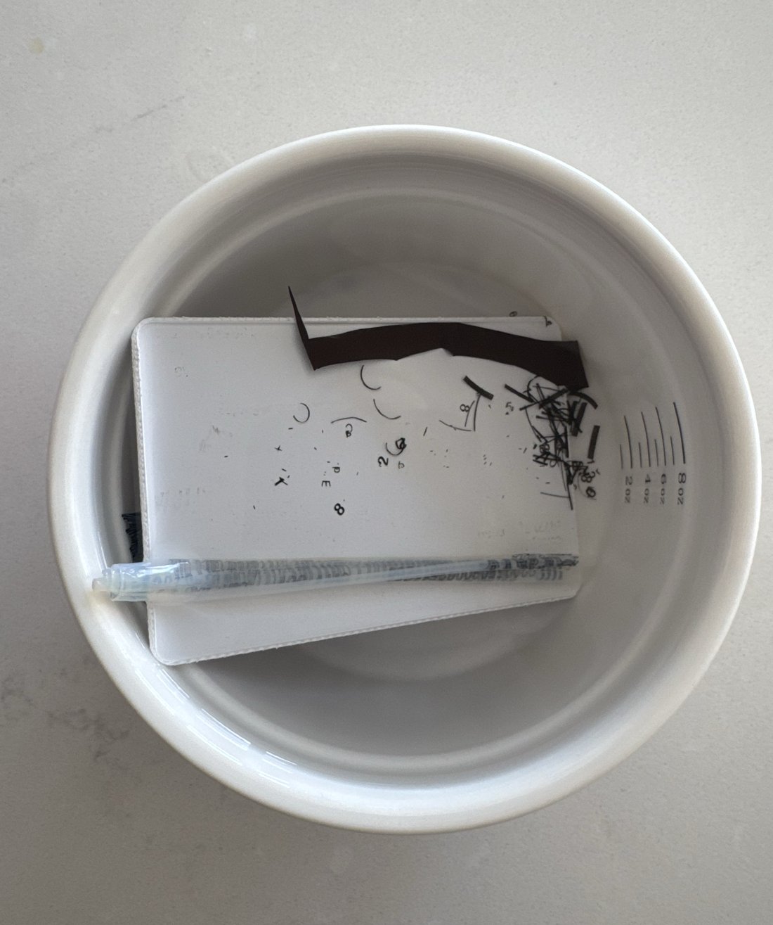 An OMNY card dissolving in a bowl of acetone, layers separating, the mag stripe peeling away