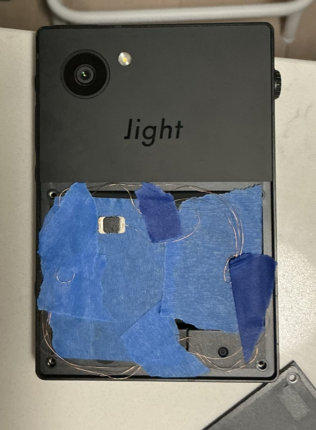 The Light Phone III's open battery compartment with the chip and antenna coil held under strips of blue painter's tape, the fine wire spread to the compartment's edges